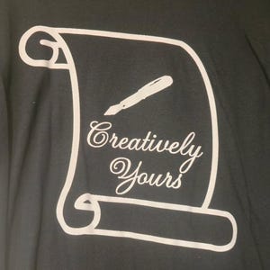 May include: Black t-shirt with a white design featuring a scroll, a pen, and the words "Creatively Yours" in a cursive font. The design is centered on the shirt.