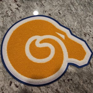 May include: A circular, textured rug featuring a stylized ram's head design. The rug has a gold-colored interior with a white outline, a blue border, and a white inner ring. The rug is placed on a speckled gray surface.