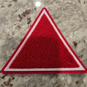 Pode incluir: Patch triangular vermelho com borda branca. O patch tem um "TM" branco no canto inferior direito.