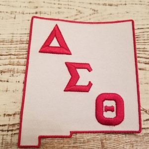 Może przedstawiać: Biała łatka z czerwonym haftowanym obramowaniem w kształcie stanu Nowy Meksyk. Łatka zawiera litery Delta Sigma Theta w czerwonym hafcie.