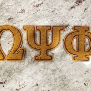 May include: Three gold Greek letters, Omega, Psi, and Phi, with a maroon outline. The letters are made of a fuzzy material and are arranged horizontally.