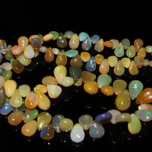 Puede incluir: Un collar de cuentas de ópalo de colores en varias formas de lágrima. Los ópalos muestran una gama de colores, incluyendo amarillo, naranja, verde, azul y blanco, con destellos iridiscentes. Las cuentas están ensartadas.