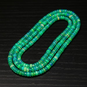 May include: A necklace composed of small, round, green beads. The beads have a slight iridescence, with hints of blue and yellow. The necklace is arranged in a looping pattern, showcasing its length and design. The background is a dark, solid color.