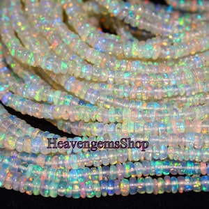 May include: A strand of white opal beads with iridescent rainbow flashes. The beads are small and round, and they are strung on a clear thread. The beads are arranged in a loose pile, and they are illuminated by a bright light. The text "HeavengemsShop" is visible in the image.