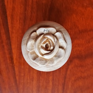 May include: A wooden knob with a carved rose design. The knob is light brown and has the letters "PTC" written on it.