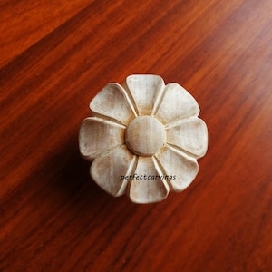 Op de afbeelding: Een gesneden houten bloem met een lichtbruine afwerking. De bloem heeft zes bloemblaadjes en een middelste cirkel. De bloem ligt op een donkerbruin houten oppervlak.