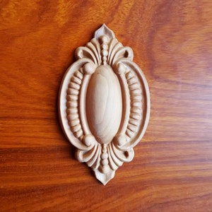 Può includere: Un accento decorativo in legno di forma ovale con intagli intricati e un centro rialzato. L'accento è di colore marrone chiaro e ha una finitura liscia.