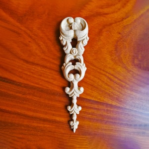 May include: A white wooden decorative accent piece with a carved, ornate design. The piece is shaped like a vertical, elongated teardrop with a series of intricate swirls and curves.