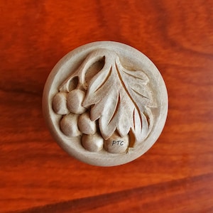 Può includere: Un pomello rotondo in legno con un design intagliato di una vite e foglie. Il pomello è dipinto di un colore marrone chiaro e ha le lettere "PTC" intagliate nella parte inferiore.