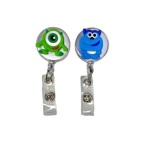 Puede incluir: Dos portagajes retráctiles con abalorios de personajes de dibujos animados. Un abalorio es un monstruo verde de un ojo y el otro es un monstruo azul.