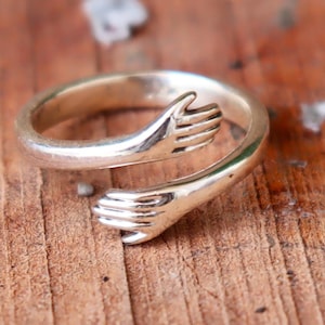 May include: A silver-colored ring with a design of two hands embracing. The ring is set against a wooden surface. The hands are detailed, with visible fingers. The ring appears to be made of metal.