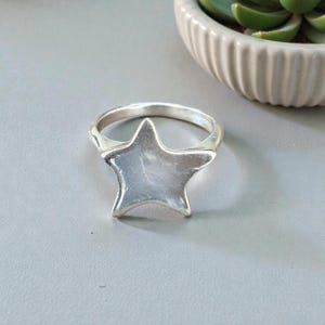 Puede incluir: Un anillo plateado con un diseño de estrella grande de cinco puntas. El anillo está sobre un fondo gris, con una maceta blanca con estrías y una planta verde en el fondo. La estrella del anillo tiene una superficie lisa y reflectante.