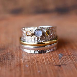 May include: A silver-tone ring with a textured band, featuring three small gemstones: two yellow and one pale blue. A gold band encircles the ring's center. The ring is displayed on a wooden surface.