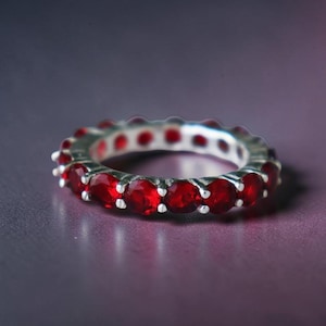 May include: A silver eternity ring featuring a continuous line of bright red gemstones. The ring's band is silver, and the gemstones are evenly spaced around the entire circumference, creating a striking and elegant appearance. Set against a purple backdrop.
