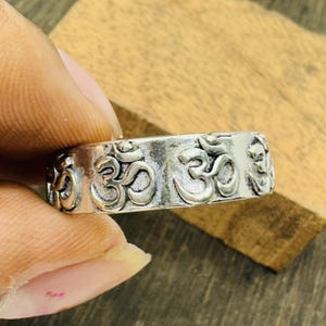 Peut inclure: Une bague en argent avec un motif en relief du symbole Om. La bague est de style jonc, avec le symbole Om répété sur toute la circonférence. La bague est tenue par les doigts d'une personne, et l'arrière-plan est une surface en bois.
