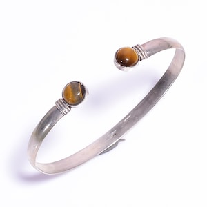 May include: A silver cuff bracelet with a tiger's eye stone at each end. The bracelet has a simple, open design with a polished finish. The tiger's eye stones are round and display a golden-brown color with natural patterns.