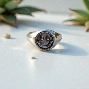May include: Silver-colored ring with a smiley face design. The ring has a rounded face with a raised smiley face in the center. The ring is made of a shiny metal and is a simple, yet stylish piece of jewelry.