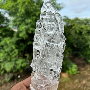May include: A clear quartz crystal sculpture of a deity, intricately carved with details and a lotus base. The sculpture is held in a hand against a blurred green background.