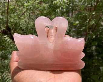 May include: A pink rose quartz sculpture of two swans with their necks intertwined, forming a heart shape. The sculpture is carved from a single piece of rose quartz and has a smooth, polished finish.