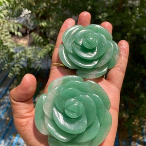 Puede incluir: Dos esculturas de rosas talladas en aventurina verde. Las rosas están detalladas con pétalos en capas y una espiral central. La piedra tiene una calidad translúcida, con variaciones en el color verde. Las esculturas se exhiben sobre un fondo borroso.