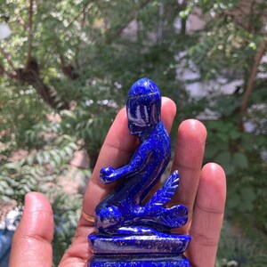 Op de afbeelding: Een lapis lazuli sculptuur van een zittende figuur met een vogelachtige staart. De diepblauwe steen heeft gouden vlekken en is gemonteerd op een rechthoekige basis. De figuur zit met uitgestrekte armen.