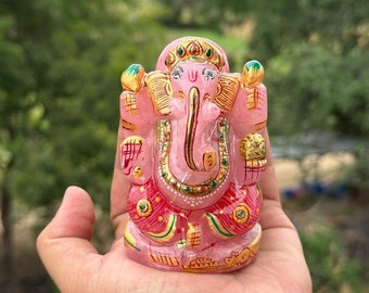 May include: A pink, hand-painted, stone Ganesha statue with gold accents. The statue is sitting on a person's hand.