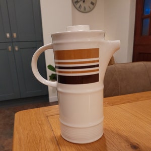 May include: White ceramic coffee pot with a brown and gold striped design. The pot has a handle and a spout.