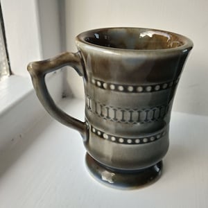 Puede incluir: Una taza de cerámica marrón y blanca con un diseño celta. La taza tiene un asa y una base.