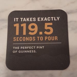 May include: A dark brown square coaster with rounded corners. The text reads "IT TAKES EXACTLY 119.5 SECONDS TO POUR THE PERFECT PINT OF GUINNESS." The coaster is on a marble surface.
