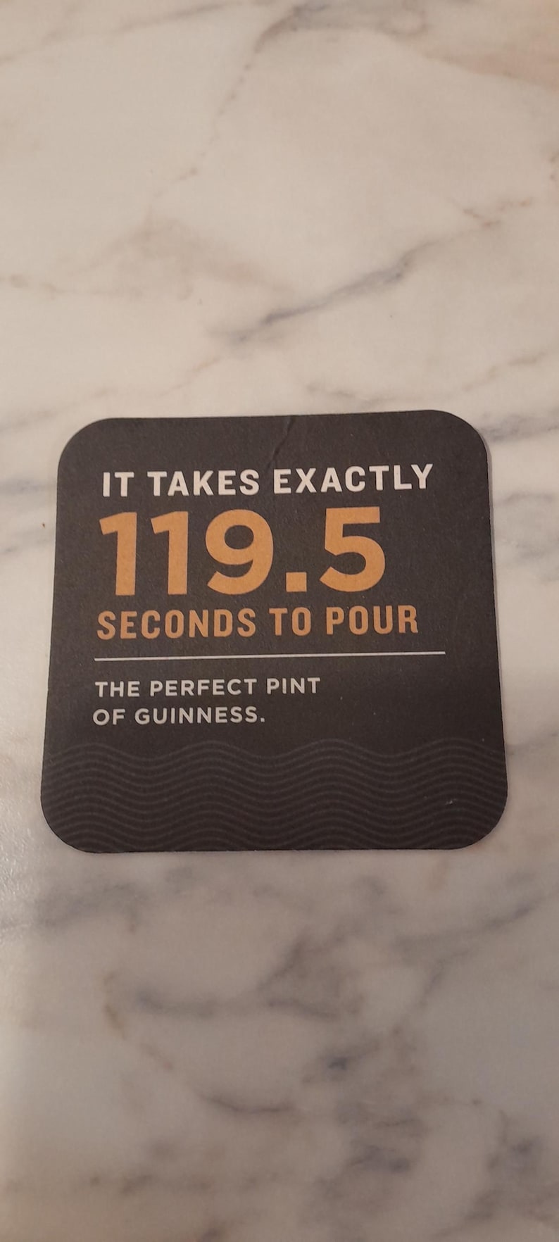 May include: A dark brown square coaster with rounded corners. The text reads "IT TAKES EXACTLY 119.5 SECONDS TO POUR THE PERFECT PINT OF GUINNESS." The coaster is on a marble surface.