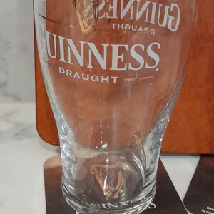 May include: A clear Guinness pint glass with the Guinness logo and the word "DRAUGHT" in white. The glass is on a coaster with the Guinness logo and text. The background is a brown surface.