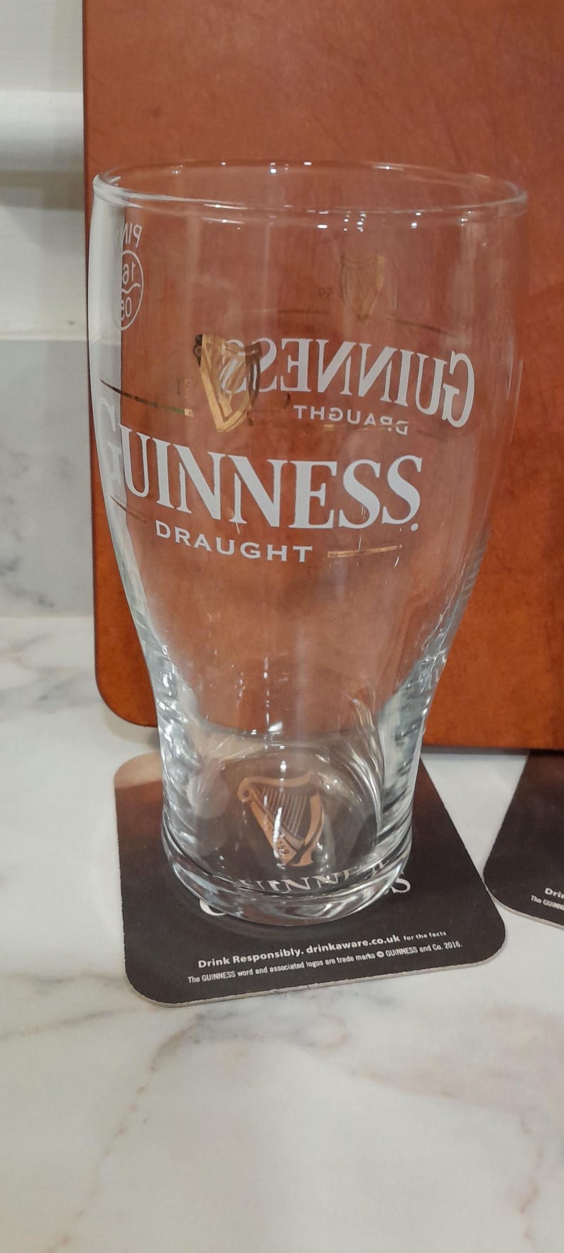 May include: A clear Guinness pint glass with the Guinness logo and the word "DRAUGHT" in white. The glass is on a coaster with the Guinness logo and text. The background is a brown surface.