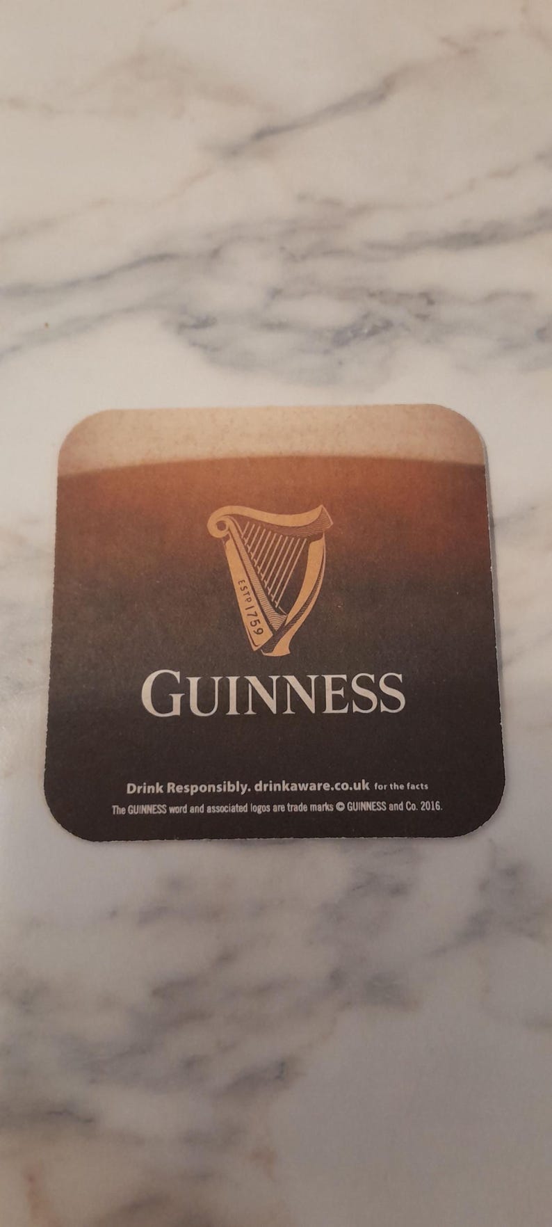 May include: Square Guinness coaster with a gradient brown and black background. The Guinness harp logo is in the center, with the word "GUINNESS" below it. Text at the bottom reads "Drink Responsibly. drinkaware.co.uk for the facts."