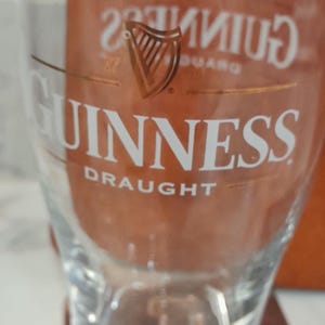 May include: A clear glass with the Guinness logo and the word "Guinness" in white lettering. The glass has a curved shape and is designed for draught beer. The logo features a harp.