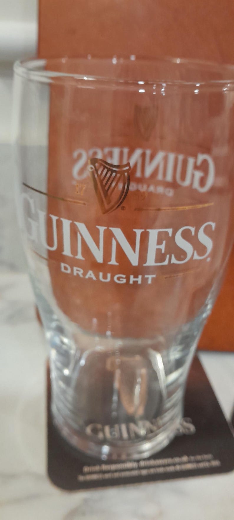 May include: A clear glass with the Guinness logo and the word "Guinness" in white lettering. The glass has a curved shape and is designed for draught beer. The logo features a harp.