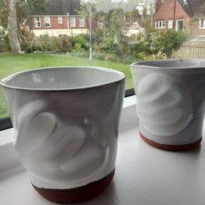 May include: Two white ceramic mugs with a textured, uneven surface. The mugs have a brown clay base.