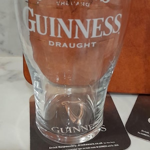 May include: A clear Guinness pint glass with the Guinness logo and "DRAUGHT" printed in white. The glass sits on a black coaster with the Guinness logo and text. A second coaster is visible.