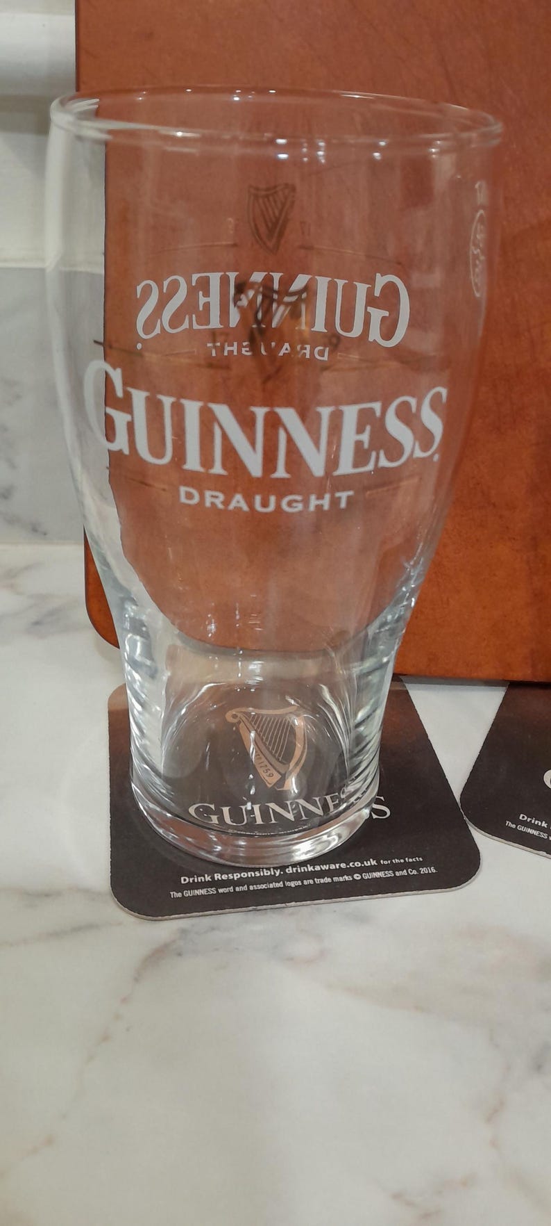 May include: A clear Guinness pint glass with the Guinness logo and "DRAUGHT" printed in white. The glass sits on a black coaster with the Guinness logo and text. A second coaster is visible.