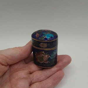 Könnte beinhalten: Eine kleine, runde, blaue und goldene Emaille-Dose mit Blumendesigns. Die Dose ist mit bunten Blumen und Blättern verziert.