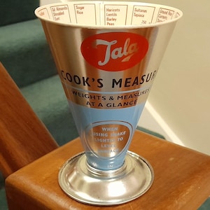 Puede incluir: Una taza medidora de cocina cónica de metal con acabado plateado y azul claro. El exterior presenta el logotipo rojo de Tala, el texto COOK'S MEASURE, WEIGHTS & MEASURES AT A GLANCE y una tabla de ingredientes impresa en el borde superior.