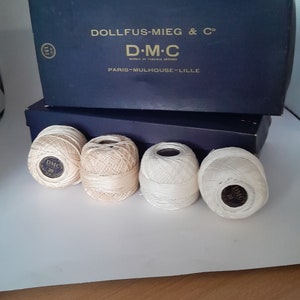 May include: Four spools of DMC embroidery floss, two are white and two are a light beige colour. The spools are sitting on a white surface. The spools have a gold label with the DMC logo and the text "DMC" in black.