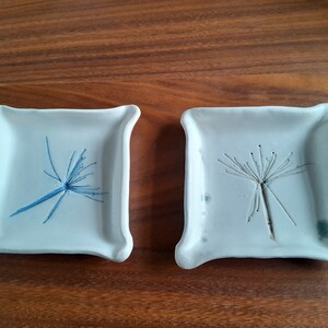 Puede incluir: Dos pequeños platos de cerámica blanca con un diseño en relieve de diente de león. Un plato tiene un diente de león azul y el otro tiene un diente de león blanco.