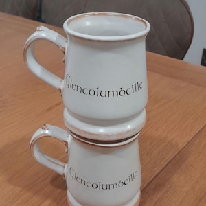 Puede incluir: Dos tazas de cerámica blanca apiladas una encima de la otra. Ambas tazas tienen un borde marrón y el texto "Glencolumcille" impreso en la parte delantera.