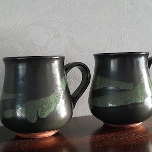 May include: Two handmade ceramic mugs. The mugs are dark green with a black rim and handle. Each mug has a green, organic-shaped design. The base of the mugs is a reddish-brown color. The mugs are on a dark brown surface.