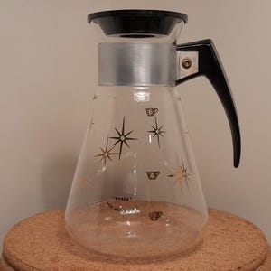 Peut inclure: Une carafe à café vintage en verre avec une poignée et un couvercle noirs, et une bande argentée. La carafe a une forme conique et est décorée d'étoiles dorées et de motifs de tasses. La carafe est posée sur une surface marron.