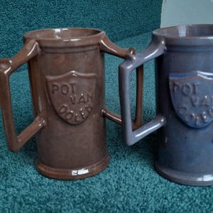 Könnte beinhalten: Zwei Keramikbecher, einer braun und einer blau, mit einzigartigen Griffen. Jeder Becher hat ein schildförmiges Emblem mit dem Text "POT VAN DOLE". Die Becher stehen auf einer türkisfarbenen Oberfläche.