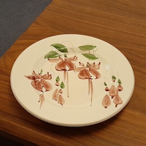 Puede incluir: Un plato de cerámica blanco con un diseño floral pintado a mano en tonos marrón, verde y crema. El plato presenta flores y hojas estilizadas. El plato es redondo y está sobre una superficie de madera.