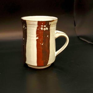 Puede incluir: Una taza de cerámica blanca con rayas marrones y rojas y manchas blancas. La taza tiene un asa.