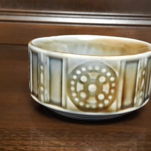 Puede incluir: Un cuenco de cerámica con forma cilíndrica. El cuenco presenta un patrón repetitivo de círculos y líneas verticales en tonos beige y verde oliva. El interior del cuenco es de color beige claro.