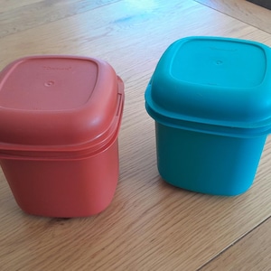 May include: Two square, lidded food storage containers. One is red, the other is teal. Both are made of plastic and sit on a wooden surface. The containers have a square shape with rounded corners and a slightly textured surface.